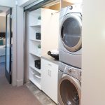 the laundry closet behind the roll up corrugated metal door is completed with cabinets and stacked washer and dryer